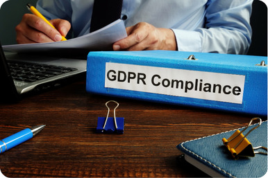 Una persona sentada en un escritorio con papeles en la mano y una computadora portátil abierta. En primer plano, hay una carpeta azul con el texto ‘GDPR Compliance’ en el lomo, destacando su importancia. Esto sugiere que el trabajo se relaciona con garantizar el cumplimiento del Reglamento General de Protección de Datos, una parte crítica de la protección de datos y la privacidad para las personas dentro de la Unión Europea.